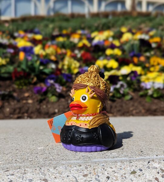 Gummiente mit traditioneller Goldhaube vor einem Blumenbeet